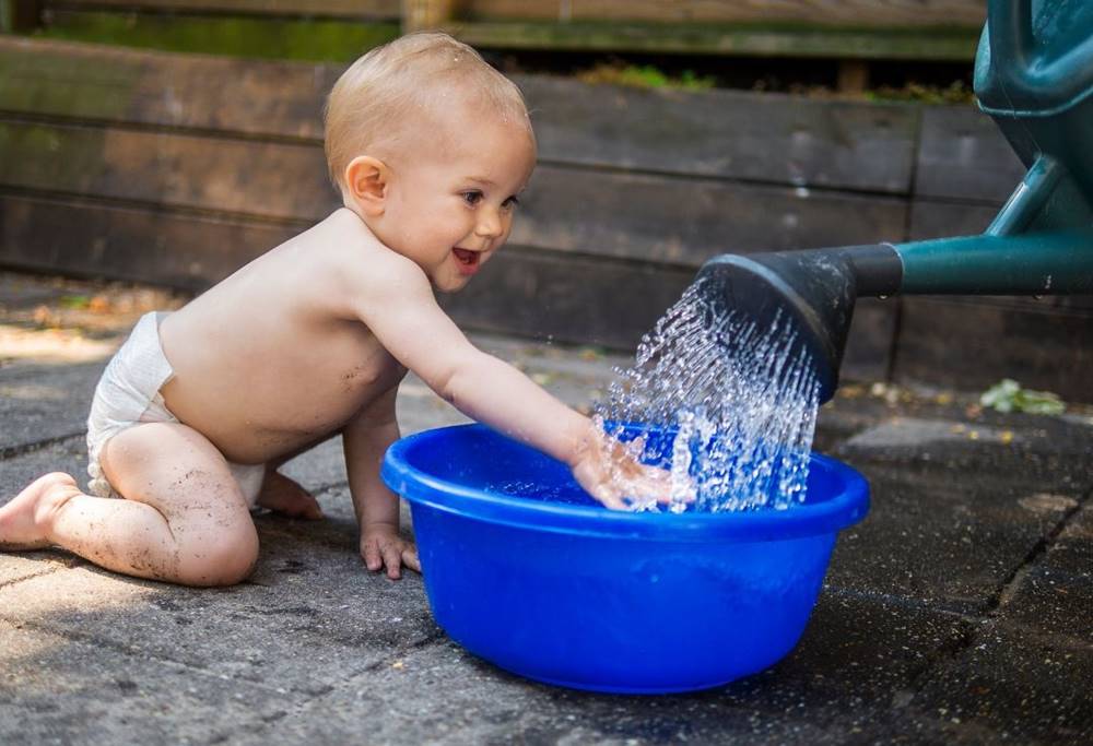 Niemowlę ciekawie wkłada rękę pod konewkę z zimną wodą – korzyści zdrowotne hartowania dziecka.